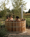 Three women relax in a wooden cedar hot tub surrounded by greenery and a silver chimney.
