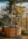 Person sitting in a wooden hot tub surrounded by trees with autumn foliage.