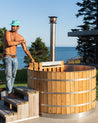 Cedar hot tub with stainless steel bands, a man stirring water, and a scenic ocean view in the background.
