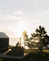 Person sitting in a wood fired hot tub by a cabin with trees and sunset in the background