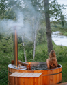 Person sitting in a wooden hot tub outdoors with trees and water in the background