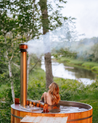 Person sitting in a wooden hot tub with steam rising, surrounded by nature.