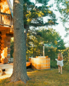Person standing near a wooden hot tub in a forest setting with a log cabin.