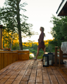 Woman standing on a wooden deck with a scenic view of trees and water.