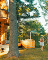 Person standing near a wooden hot tub outdoors with a log cabin and trees in the background