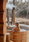Woman in a wooden hot tub with a view of a forest and stream.