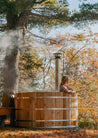 Person sitting in a wooden hot tub surrounded by trees with autumn foliage