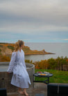 Person in a white robe standing by a hot tub with a scenic view of the ocean and cliffs.