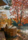 Person in a wooden hot tub surrounded by autumn trees and a lake.