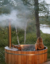 Person sitting in a wooden hot tub outdoors with mist rising from it, surrounded by nature.